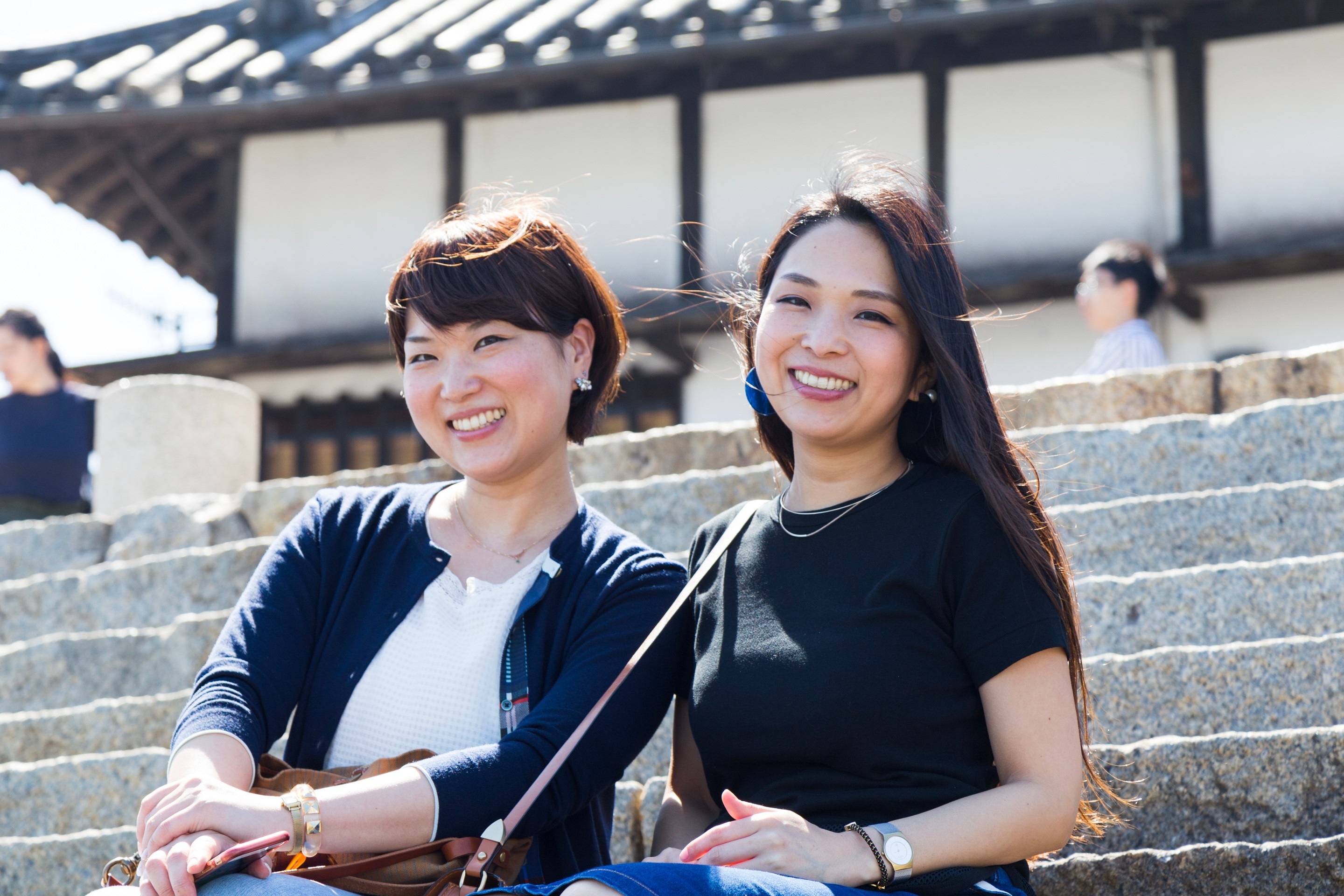 Two young smiling Japanese women sitting on stone steps in Tomonoura, Hiroshima Prefecture.