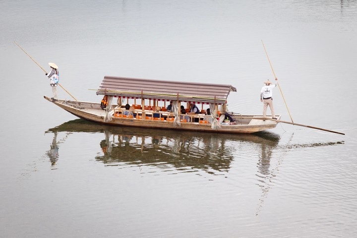 Two Japanese punters, wearing traditional clothing and wide conical hats, punting a tourist boat across the Nishiki River in Iwakuni City, Yamaguchi Prefecture.