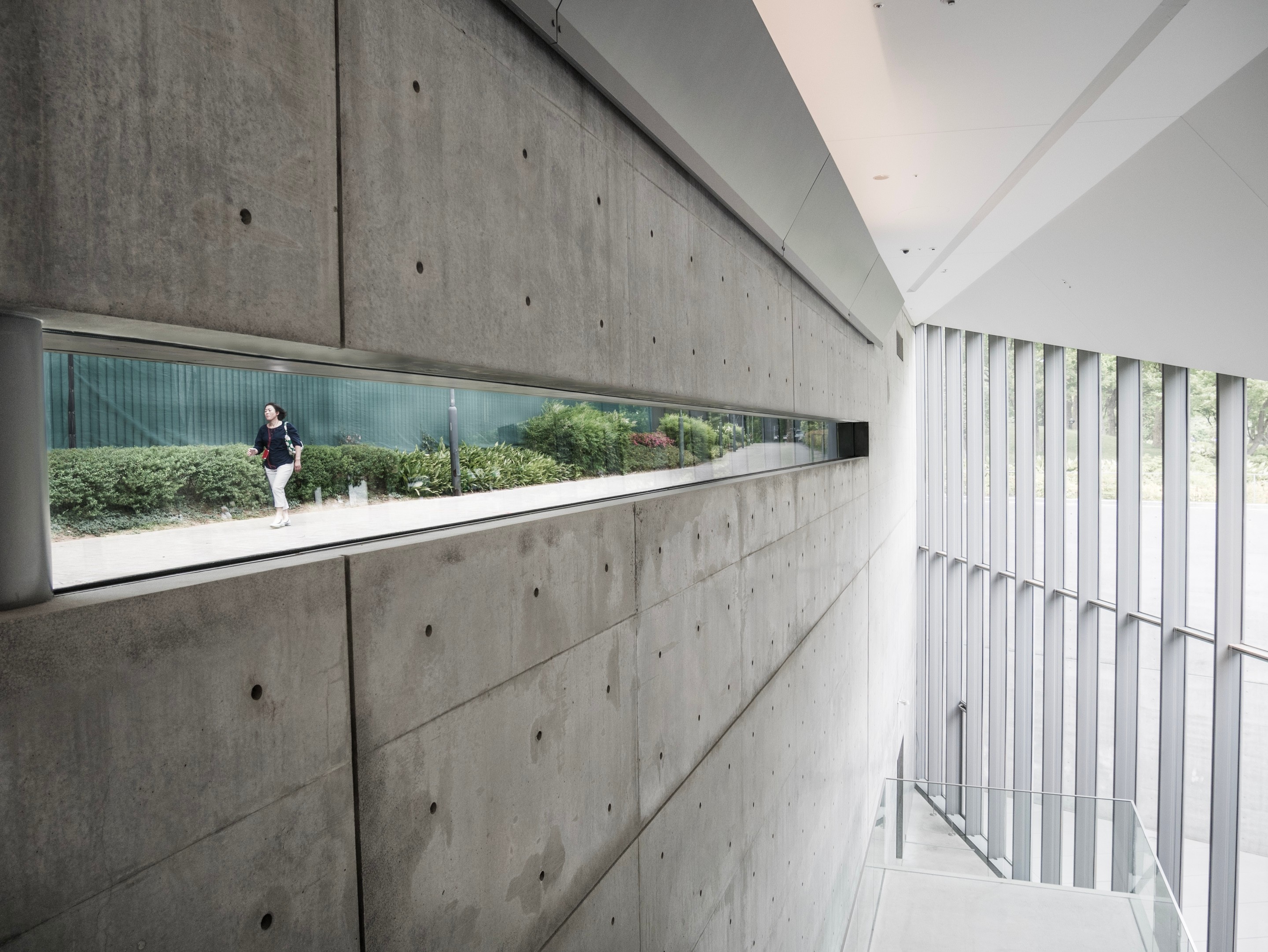 A woman can been seen walking to work through the long and narrow window, which when juxtaposed to the concrete wall appears to be a surreal fish tank, at 21_21 Design Sight Gallery — Roppongi, Tokyo, Japan