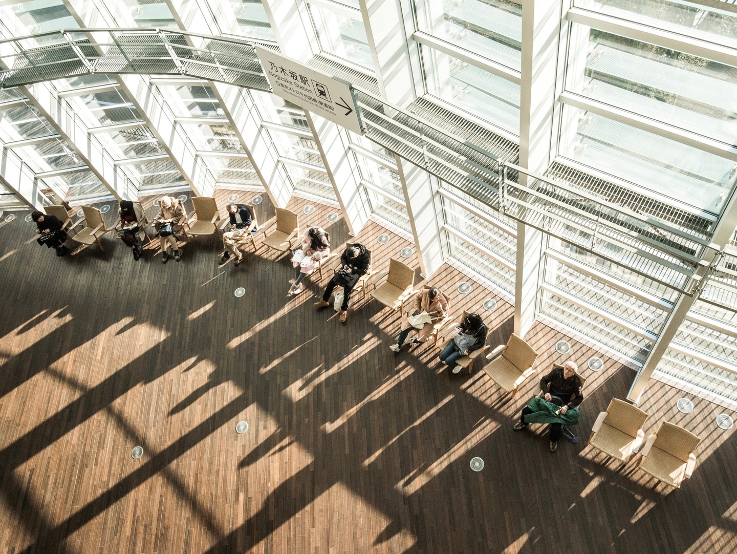 Looking down to the main atrium at The National Art Center Tokyo. The huge walls of windows cast long shadows across the wooden floor. Chairs full of resting patrons follow the contors of the wall — Roppongi, Tokyo, Japan