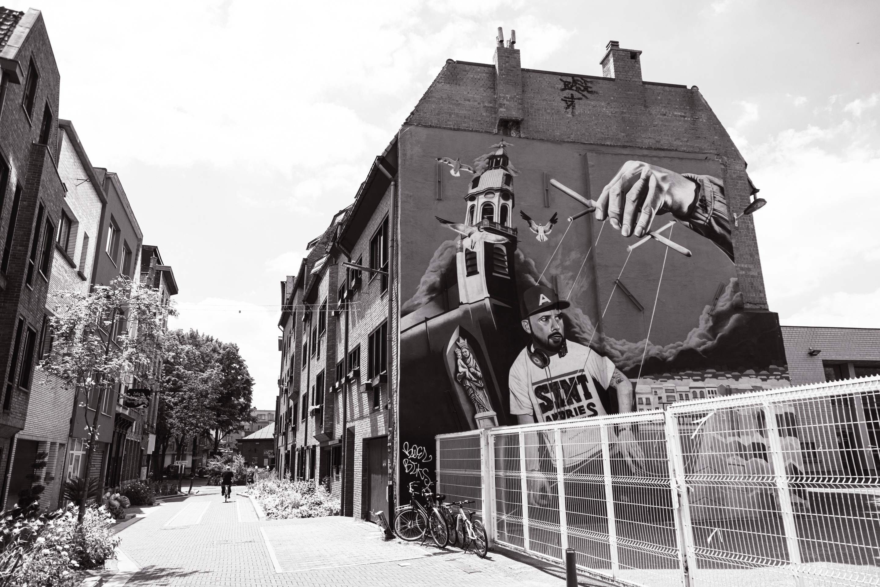 A large mural on the side of a terraced house, which forms one wall within the grounds of a school playground — depicts a young man being controlled as if a puppet, along with religious inconography — Antwerp City, Belgium