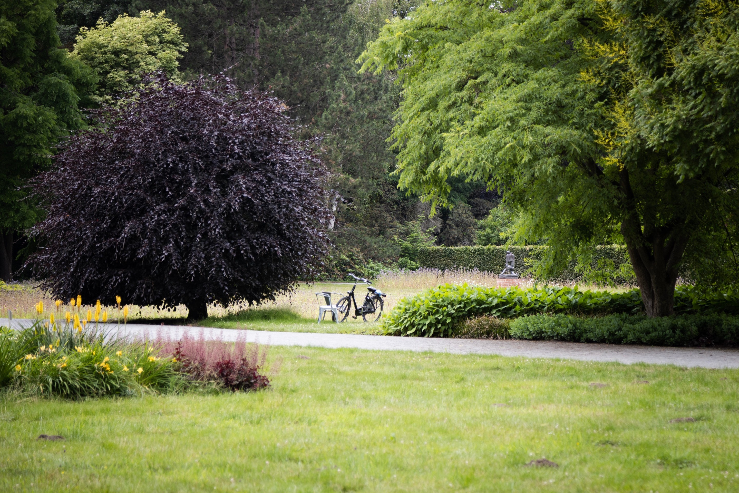 An old bicycle standing in park between two metal chairs. Behind, to the right is a metal scurlture. The scene is green a lush with trees and flowers — Middelheim Museum, Antwerp, Belgium