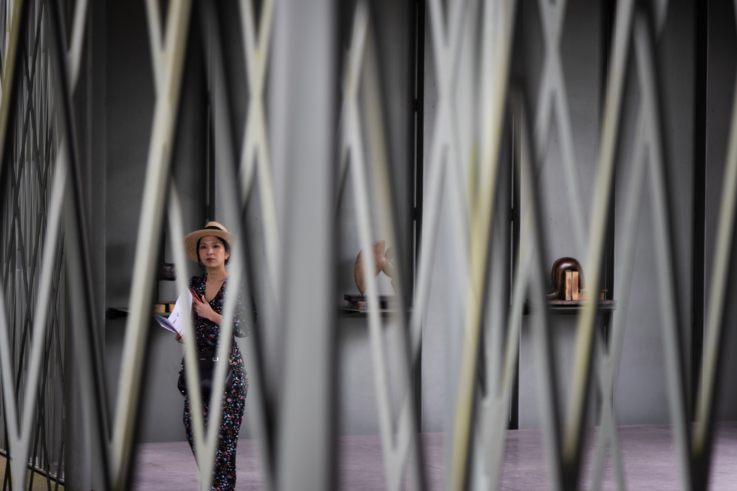 Looking through an iron lattice screen a woman looks to the camera whilst she browses through an architectural open-air sculpture gallery with works by Camille Henrot on display — Middelheim Museum, Antwerp, Belgium