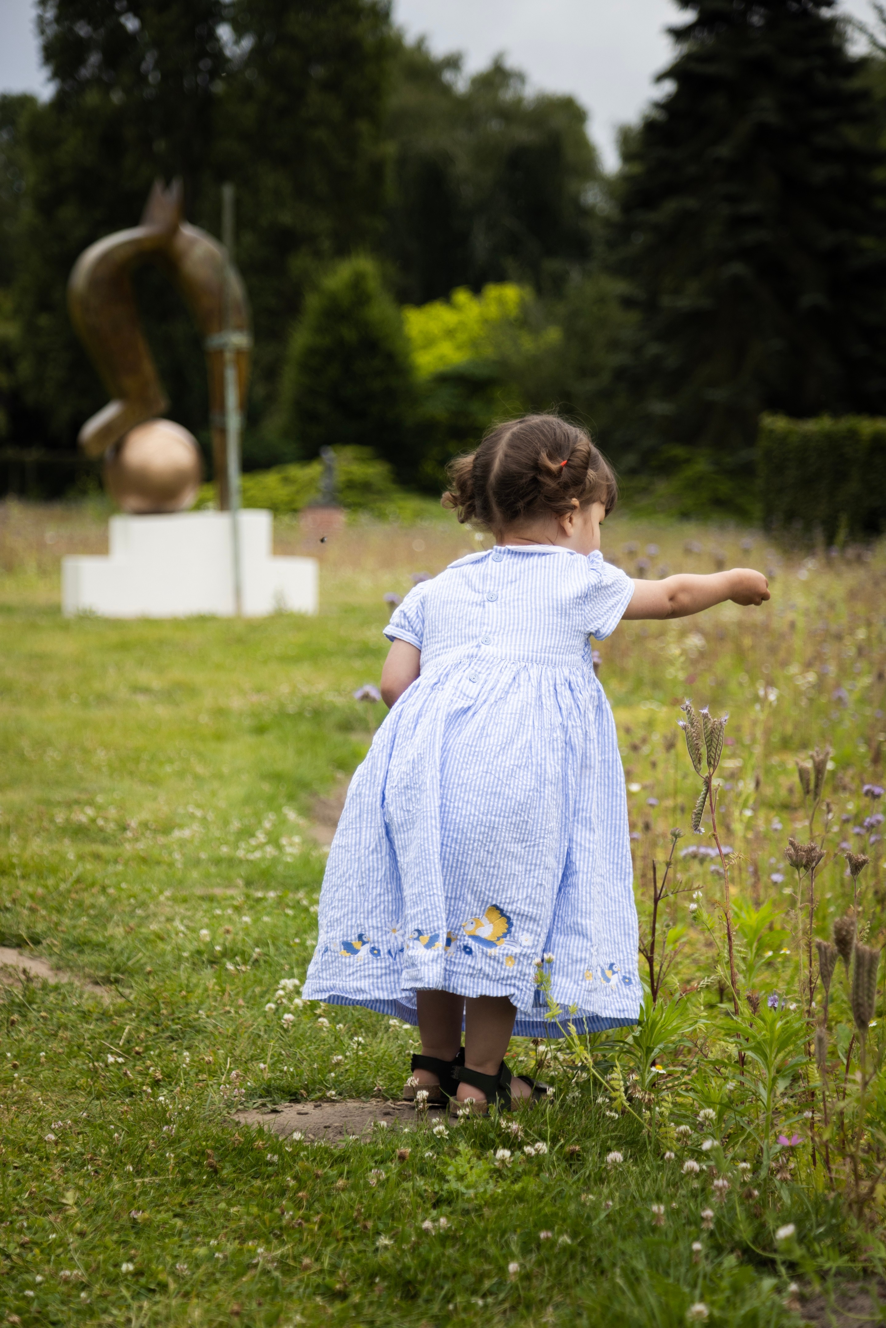 A young girl wearing a blue striped dress stands in a wildflower garden with a bronze sculture in the background. She tries to attract a butterfly by offering it some flowers — Middelheim Museum, Antwerp, Belgium