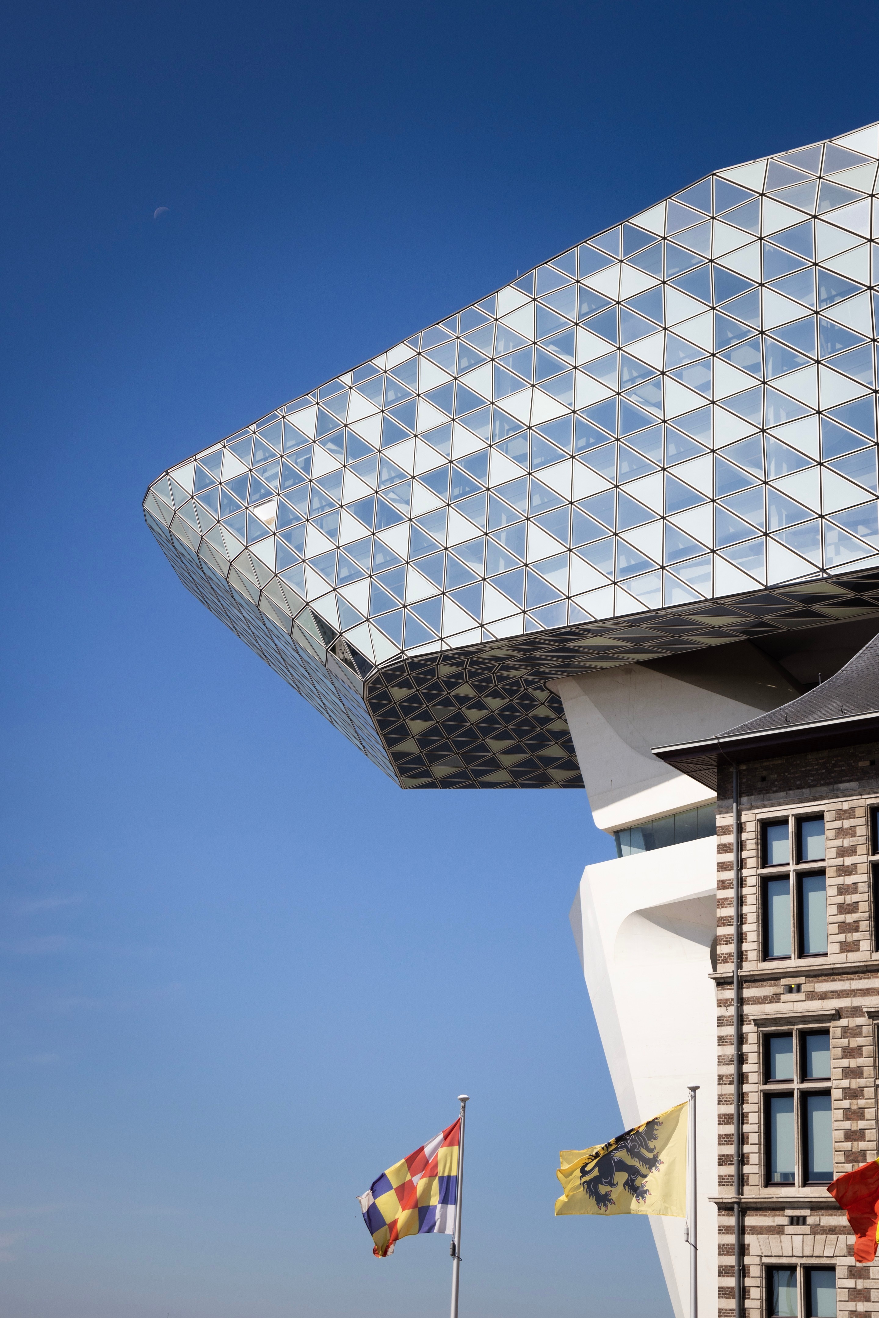 Front east side elevation of the Port House by Zaha Hadid, with a small section of the original firestation building visible. In the foreground are three municiple flags — Antwerp, Belgium