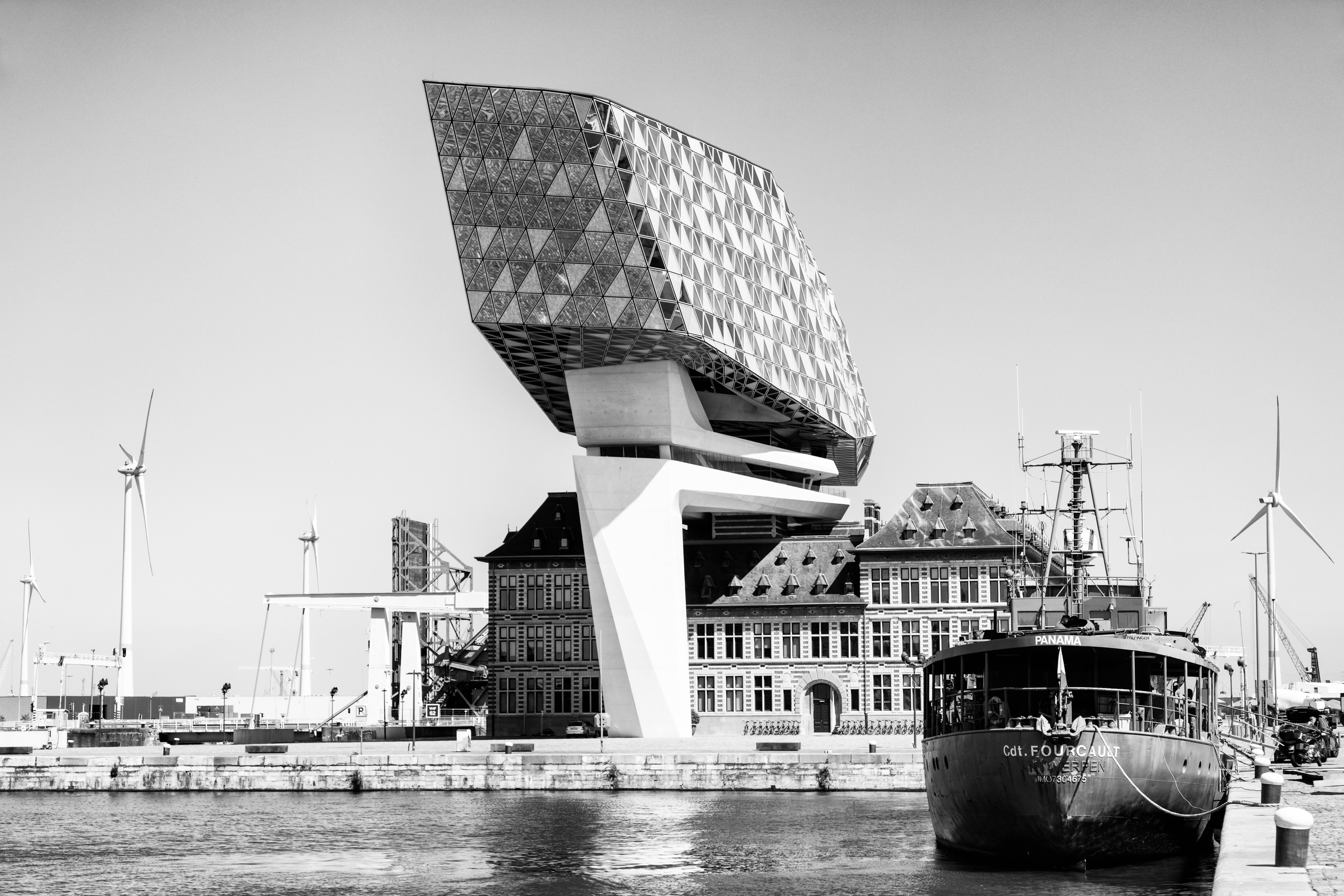Dramatic high contrast monochrome photo of the south elevation of the Port House by Zaha Hadid in the background, with the harbour water and a moored iron ship in the forground left — Antwerp, Belgium