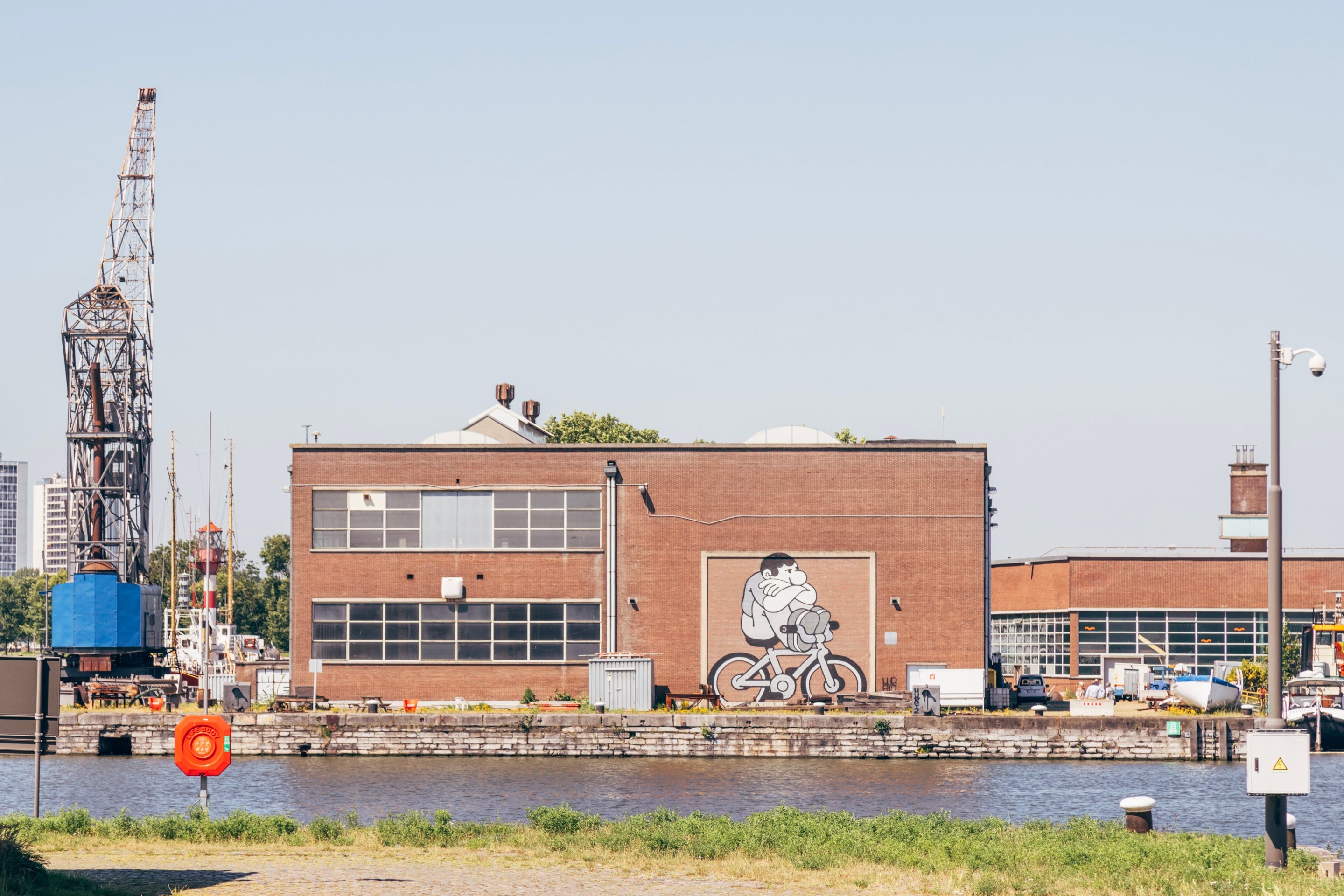 Street art of an illustrative cyclist that's given up cycling (hands folded, and feet on handlebars) on the side of a brick warehouse building, with port water in th foreground — Antwerp, Belgium