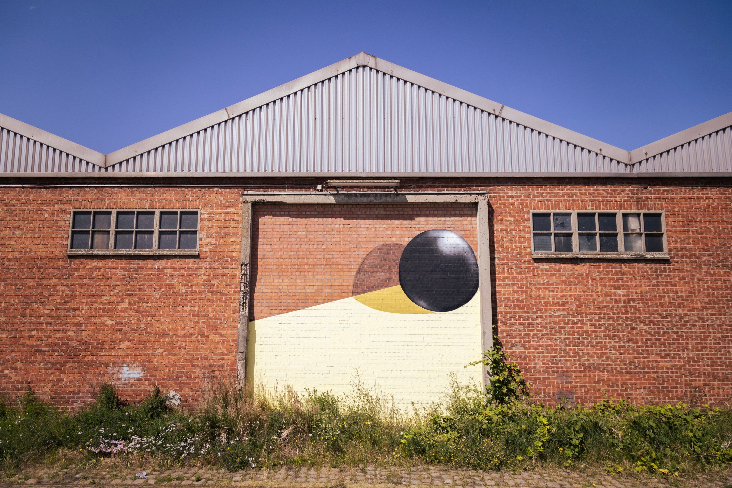 Street art of a ball, it's shadow and a yellow polygon framed by crumbling concrete on the side of a brick warehouse building — Port area of Antwerp, Belgium