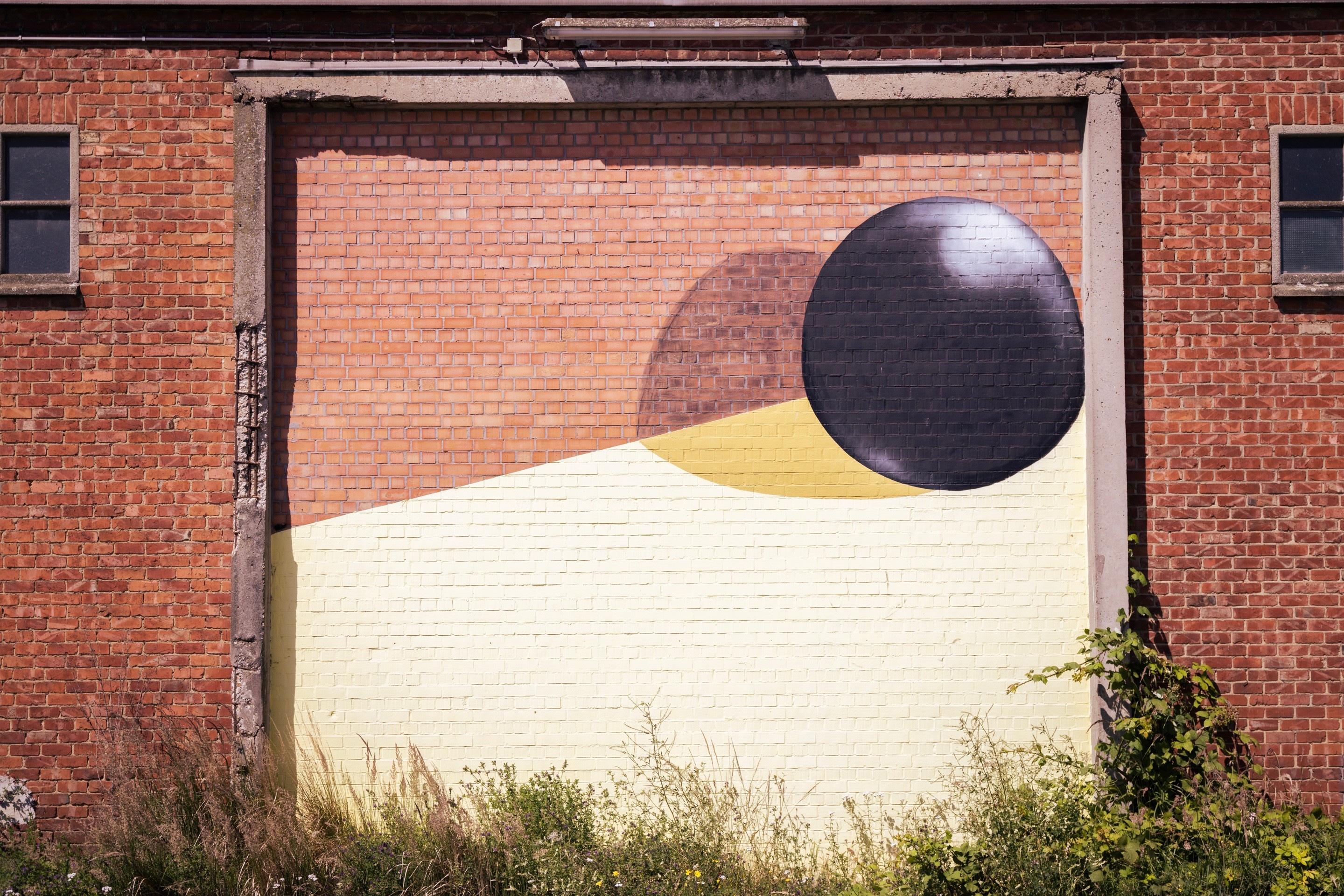 Street art of a ball, it's shadow and a yellow polygon framed by crumbling concrete on the side of a brick warehouse building — Port area of Antwerp, Belgium