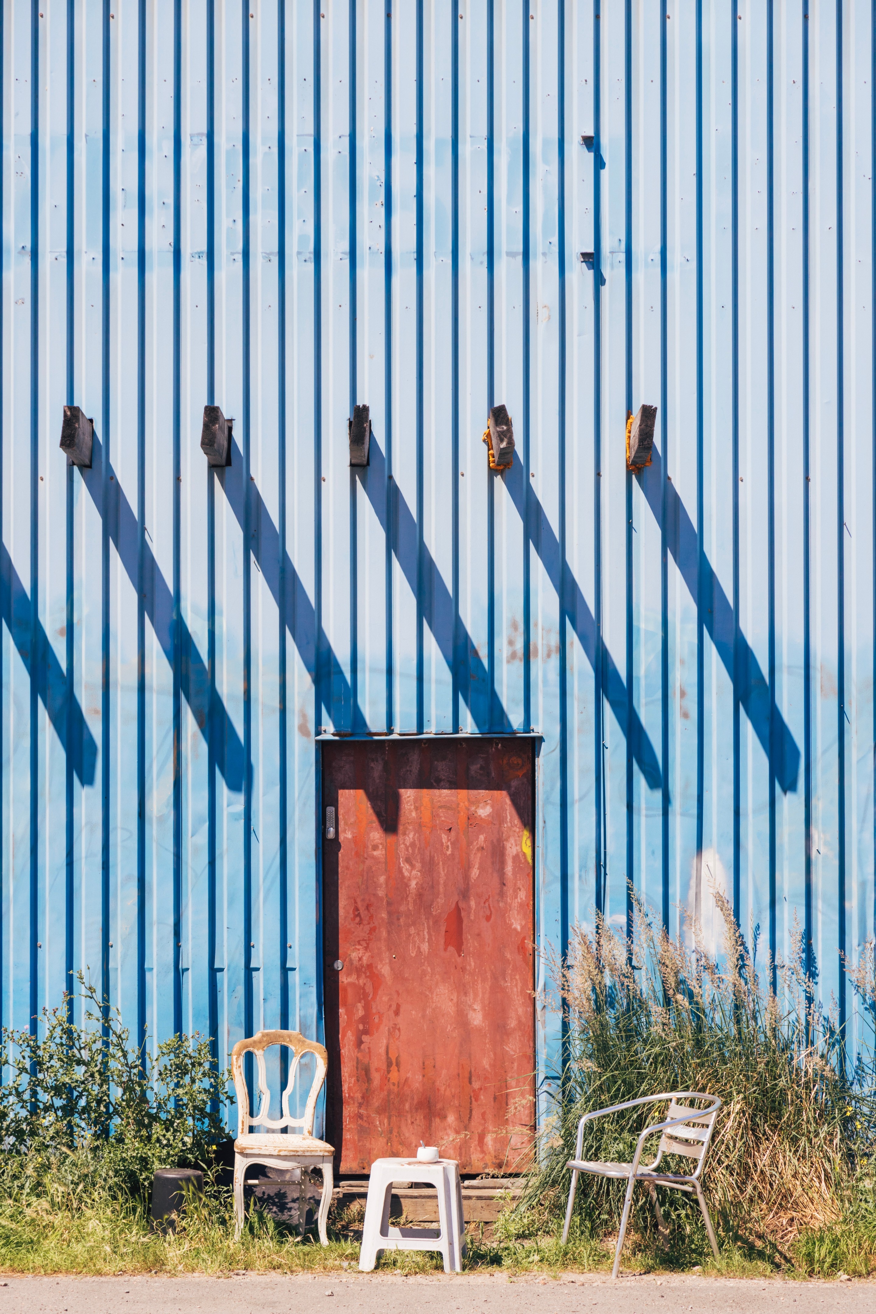 Two chairs and a stool serve as breakout smoking area behind an old faded blue warehouse building; sunlight casts a series of repeating shaddows from beams that protrude through the building's metal wall — Port area of Antwerp, Belgium