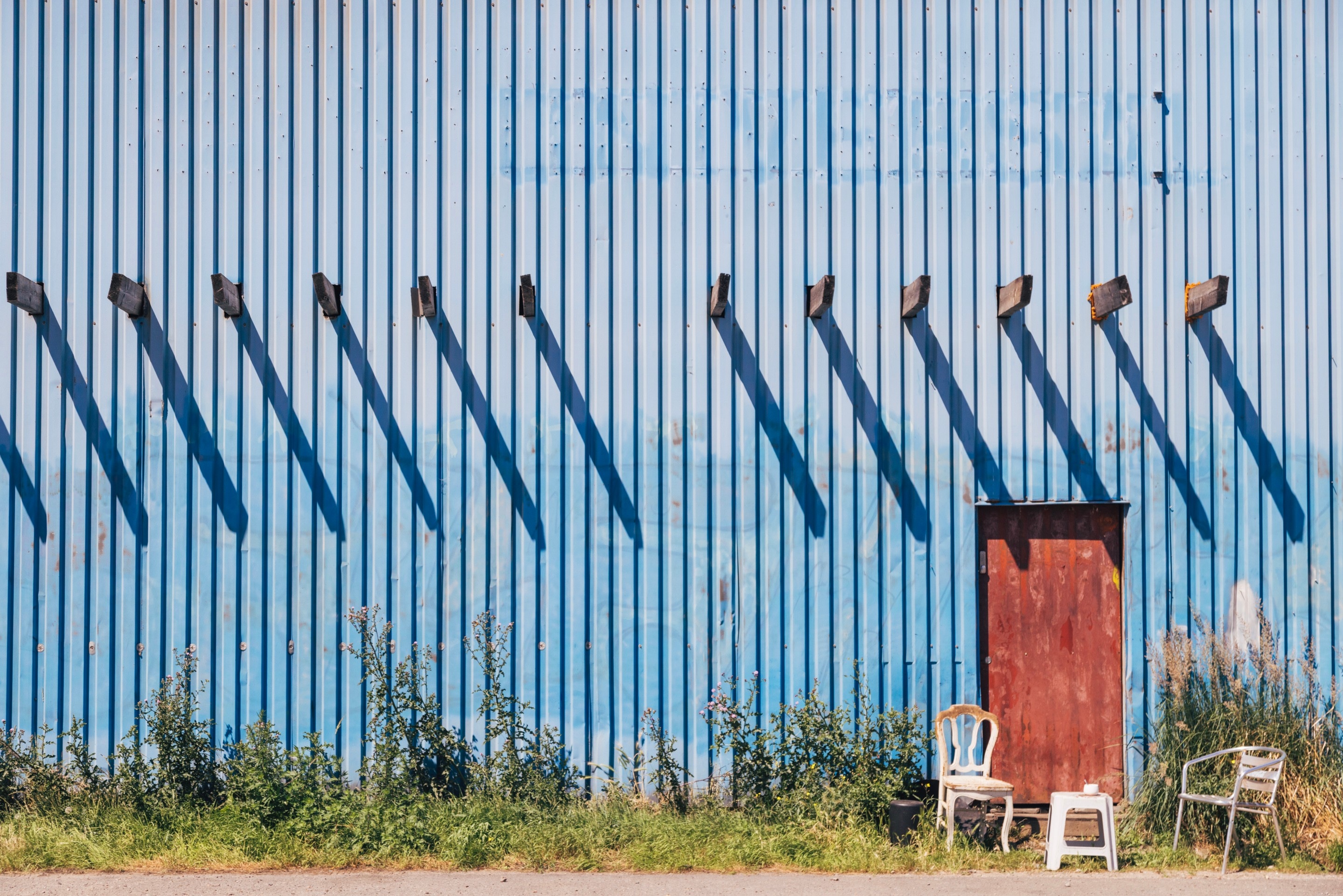 Two chairs and a stool serve as breakout smoking area behind an old faded blue warehouse building; sunlight casts a series of repeating shaddows from beams that protrude through the building's metal wall — Port area of Antwerp, Belgium