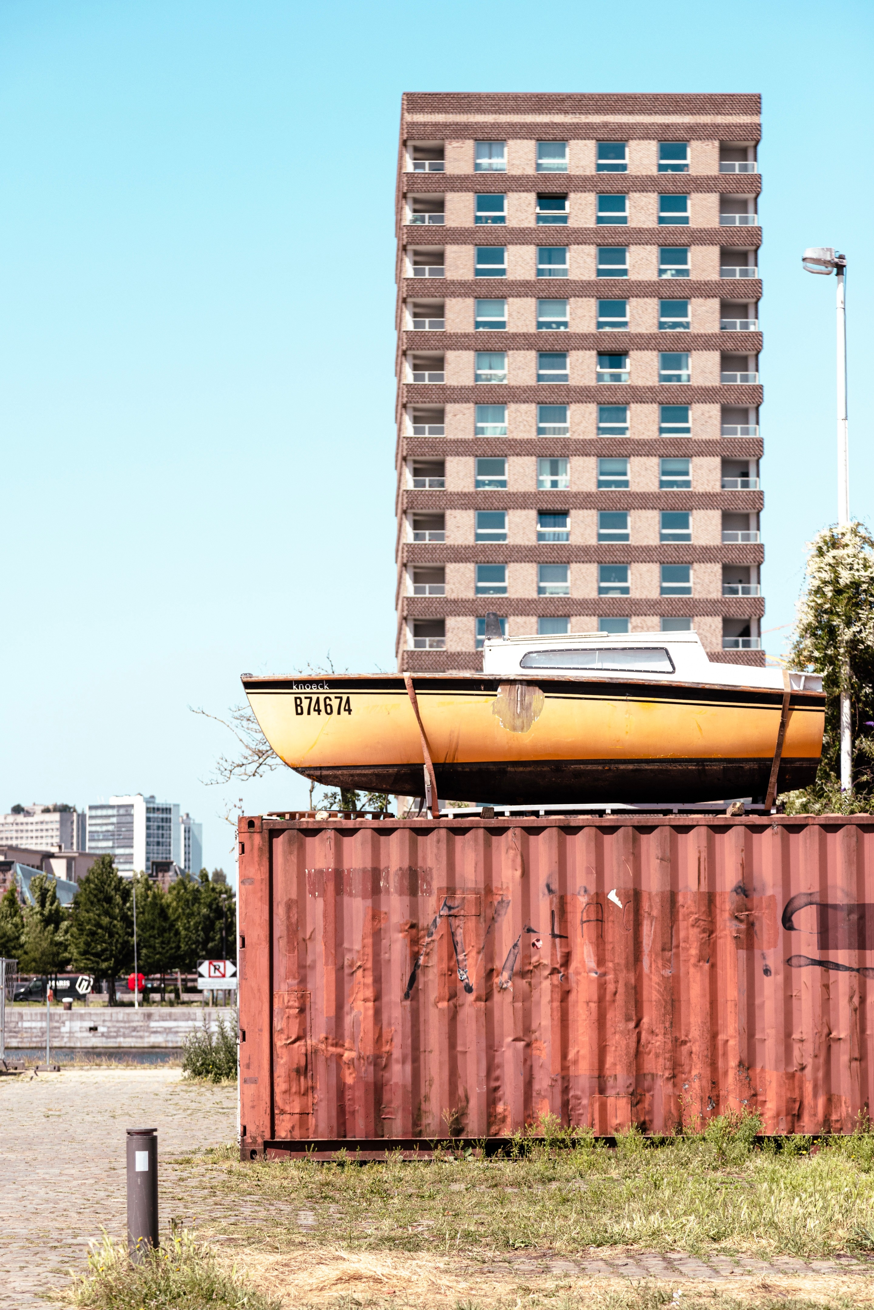 A small fibreglass boat with cabin rest atop a rusting freight container. In the background is a brutalist high rise block of flats — Port area, Antwerp, Belgium