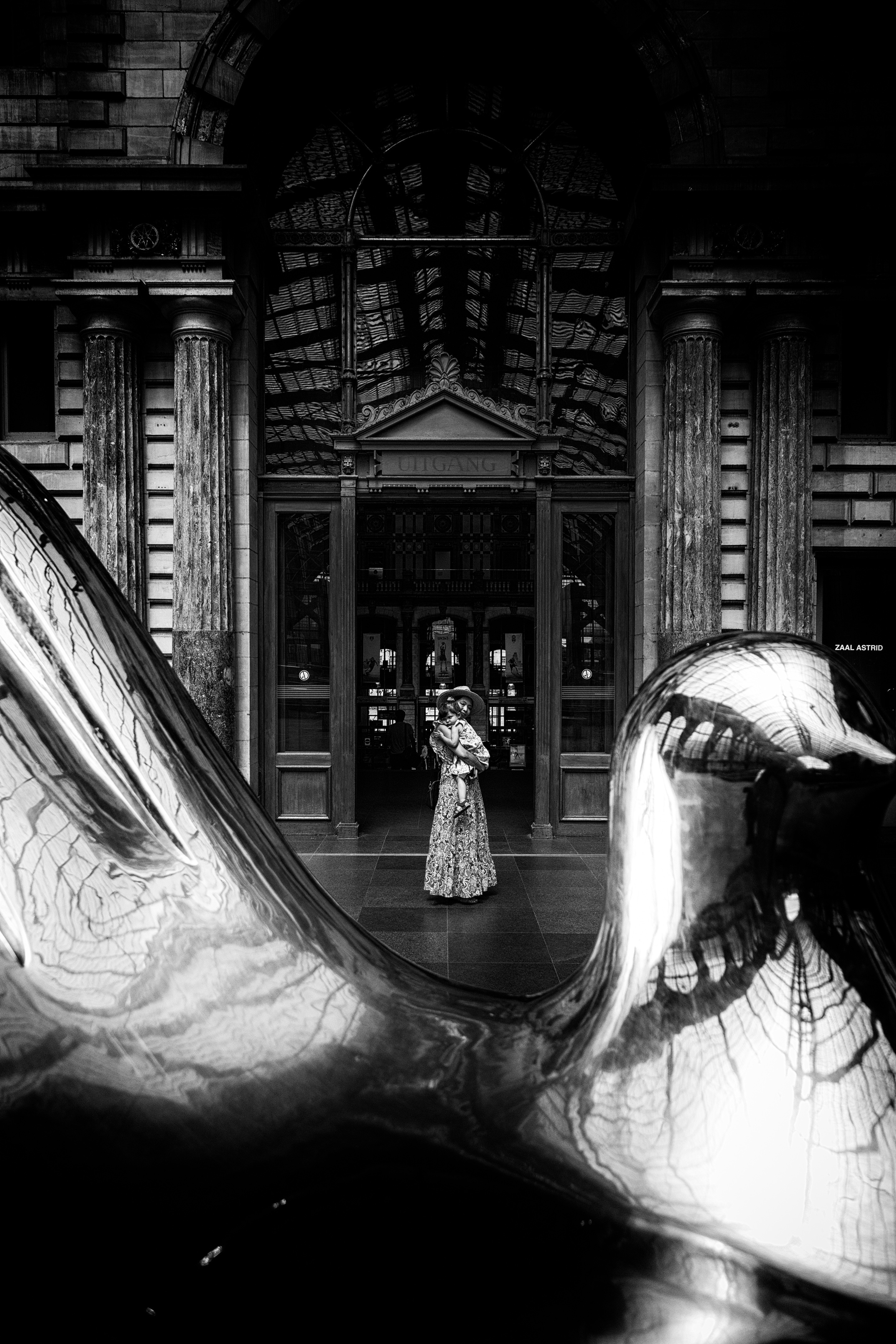 A dramatic black and white photo of a mother hugging her daughter in the doorway from the main atrium of the to the platforms, they are framed by a huge large polished metal sculture of an open hand — Antwerp Central Station, Antwerp, Belgium
