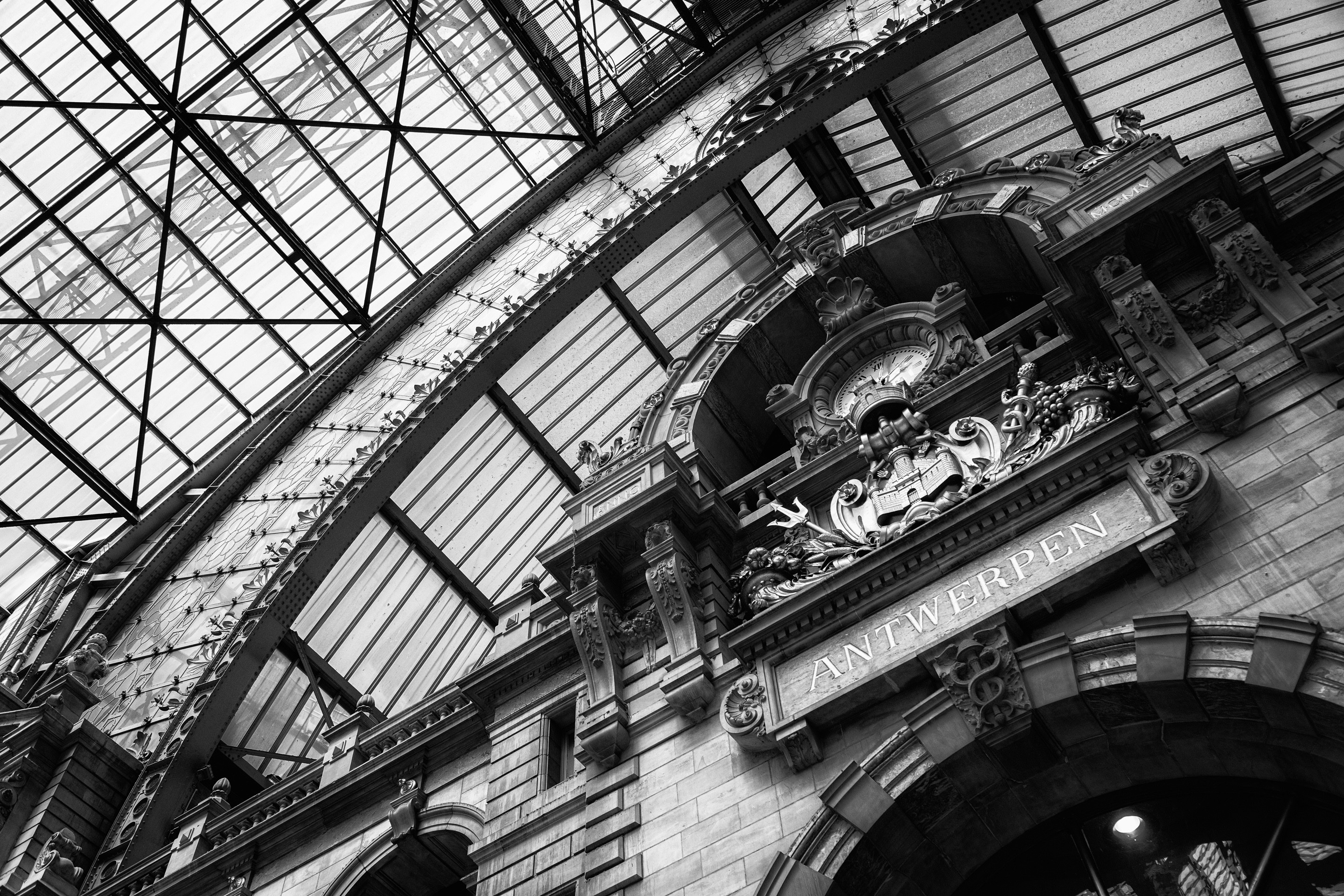A dramatic black and white photo of the main station clock, the Antwerpen coat of arms, and the impressive ironwork of the station roof — Antwerp Central Statio, Antwerp, Belgium
