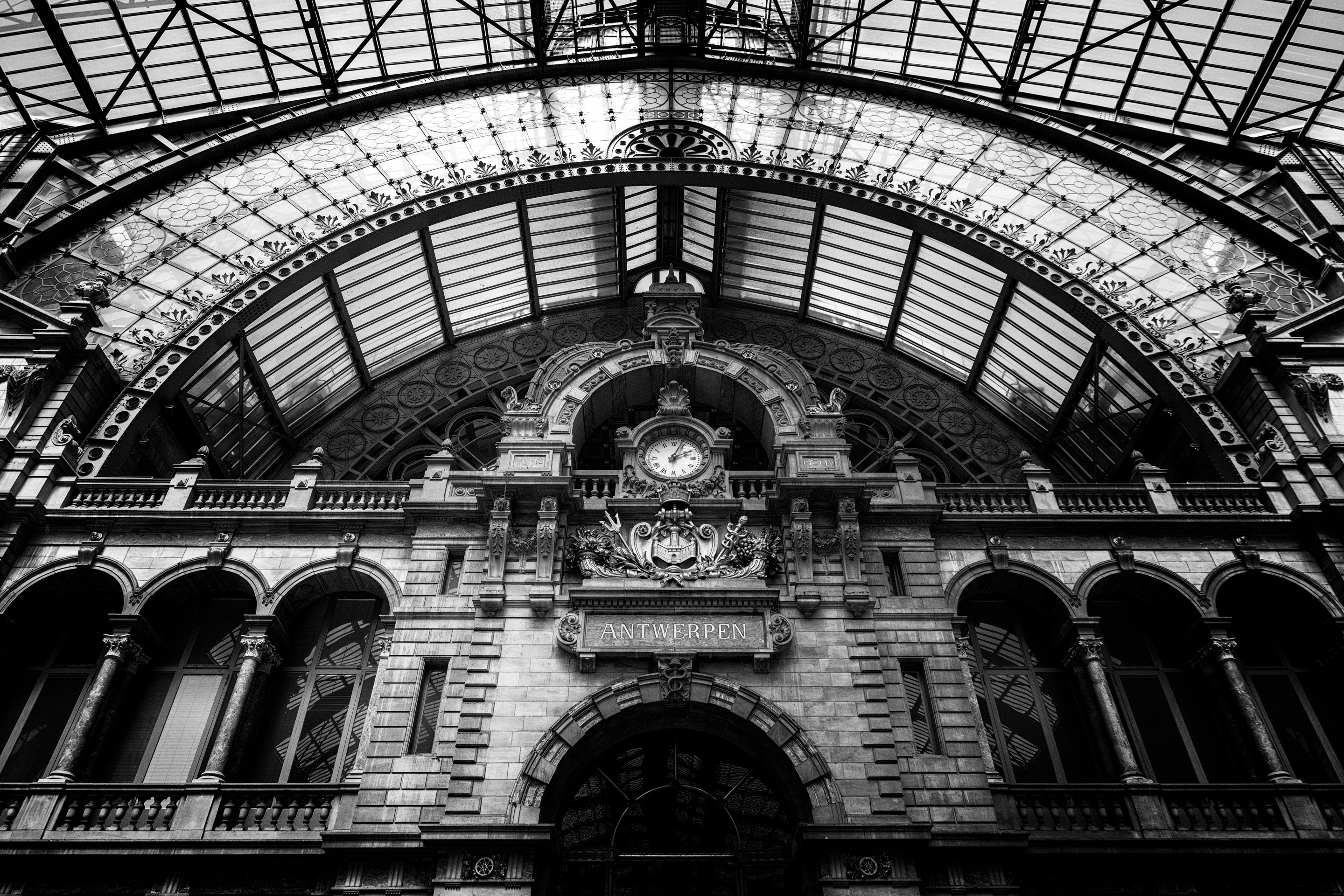 A dramatic black and white photo of the main station clock, the Antwerpen coat of arms, and the impressive ironwork of the station roof — Antwerp Central Statio, Antwerp, Belgium