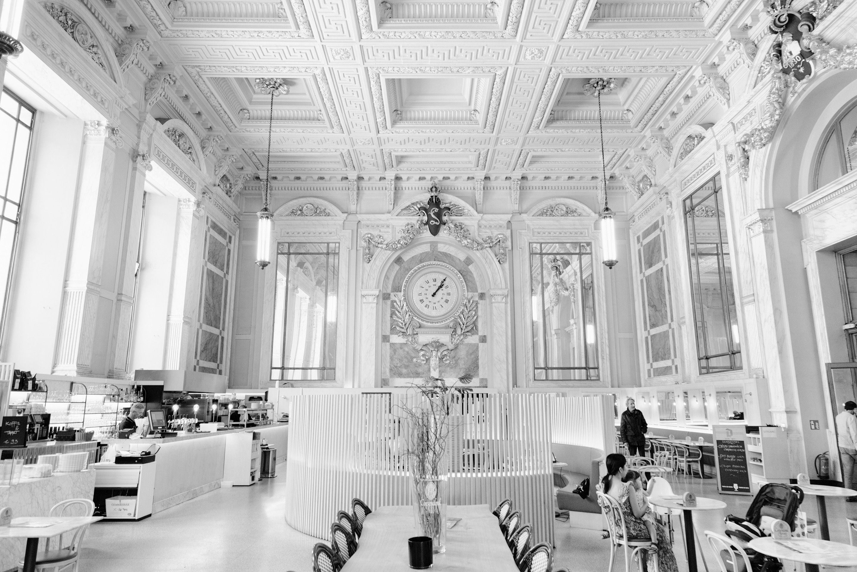 A light and airy black and white photo of the main cafe at Antwerp Central Station, the photos shows the impessive neo-Baroque and art nouveau ceiliings, clock and large mirrors that surround the cafe — Antwerp, Belgium
