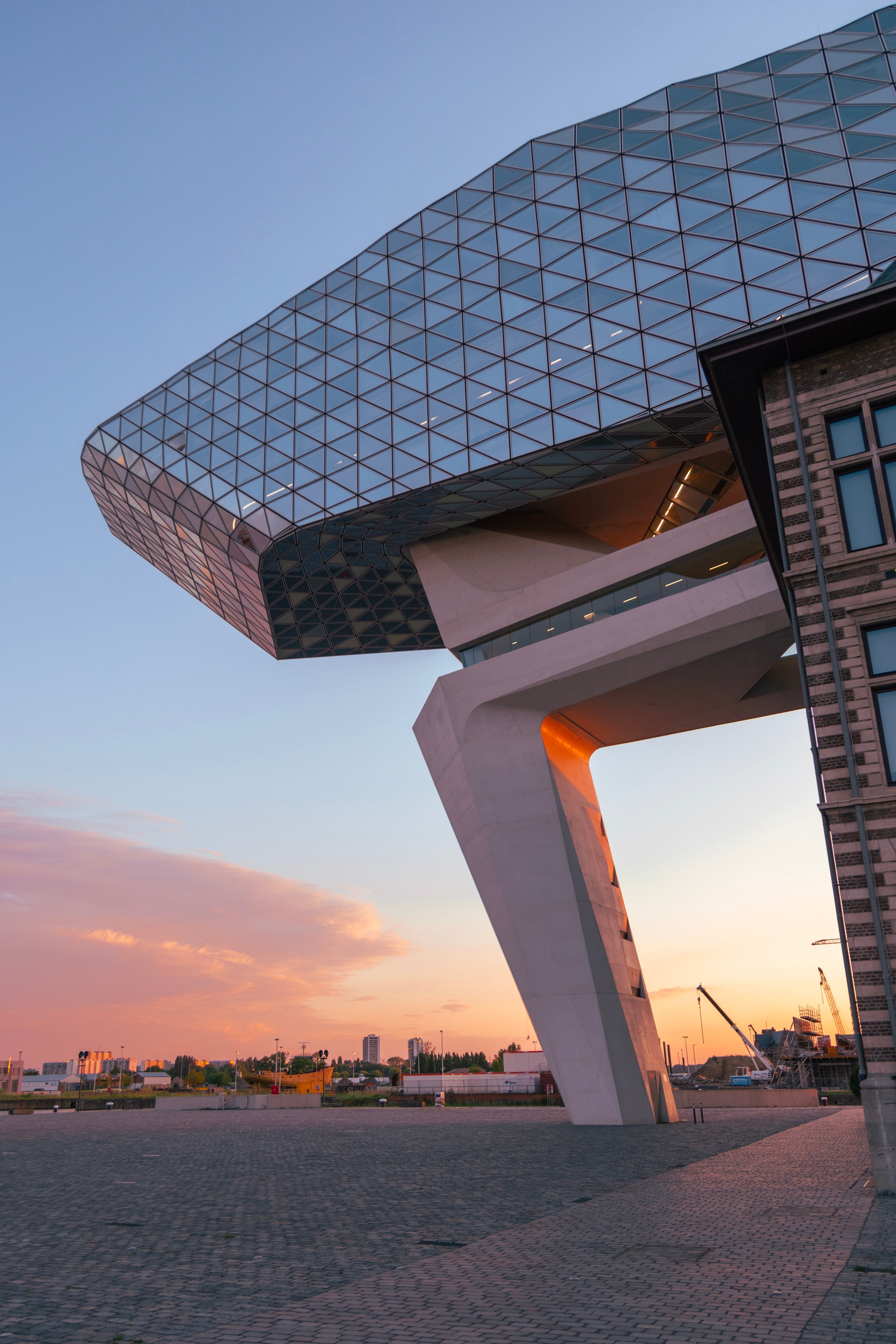 Southeastern elevation of The Port House  by Zaha Hadid during golden hour, with golden orange sunlight illuminating the concrete cantilevered support column — Antwerp, Belgium