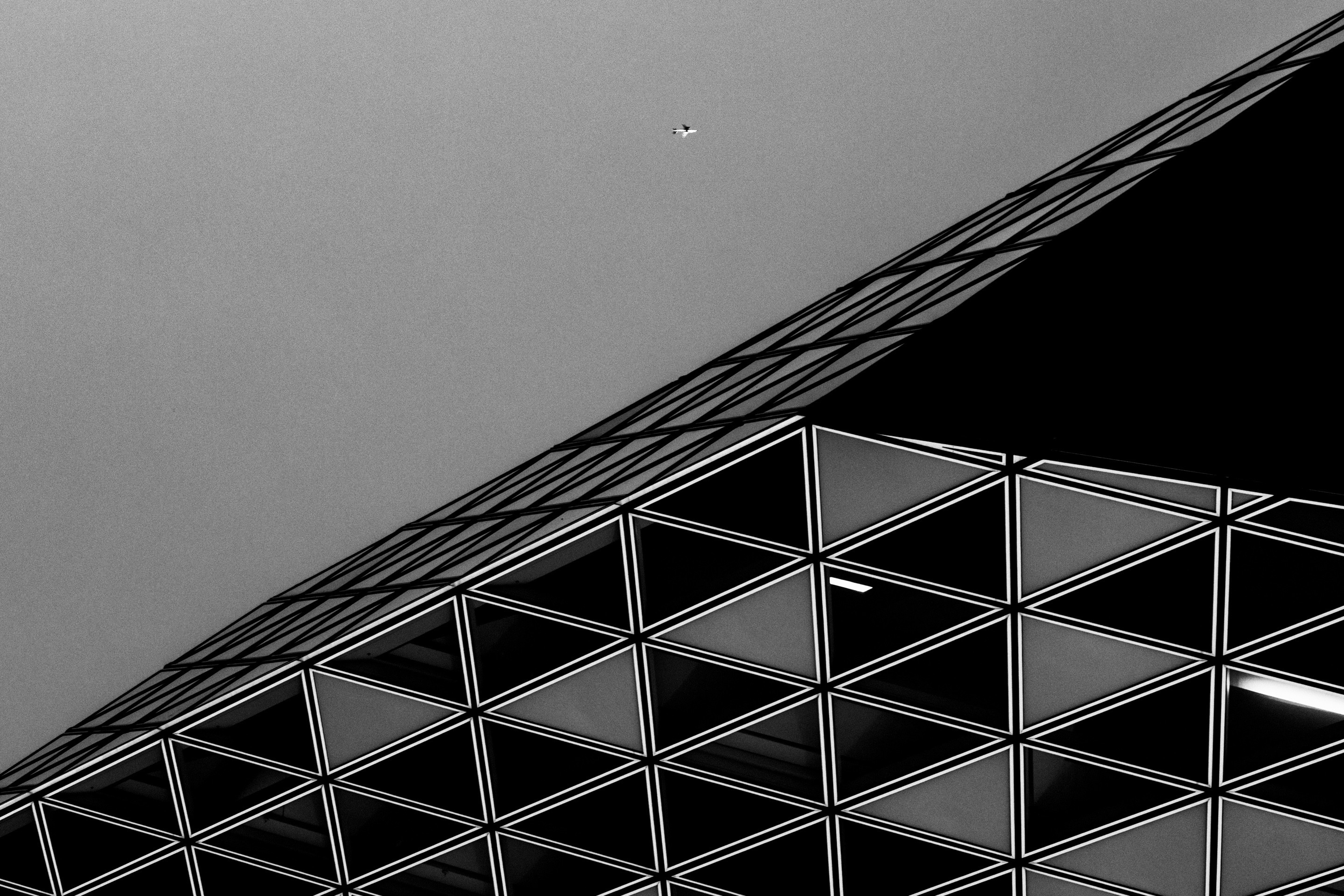 Dramatic high contrast close-up section from the glass extension of The Port House by Zaha Hadid; at the top of the frame is a aeroplan mid-flight — Antwerp, Belgium