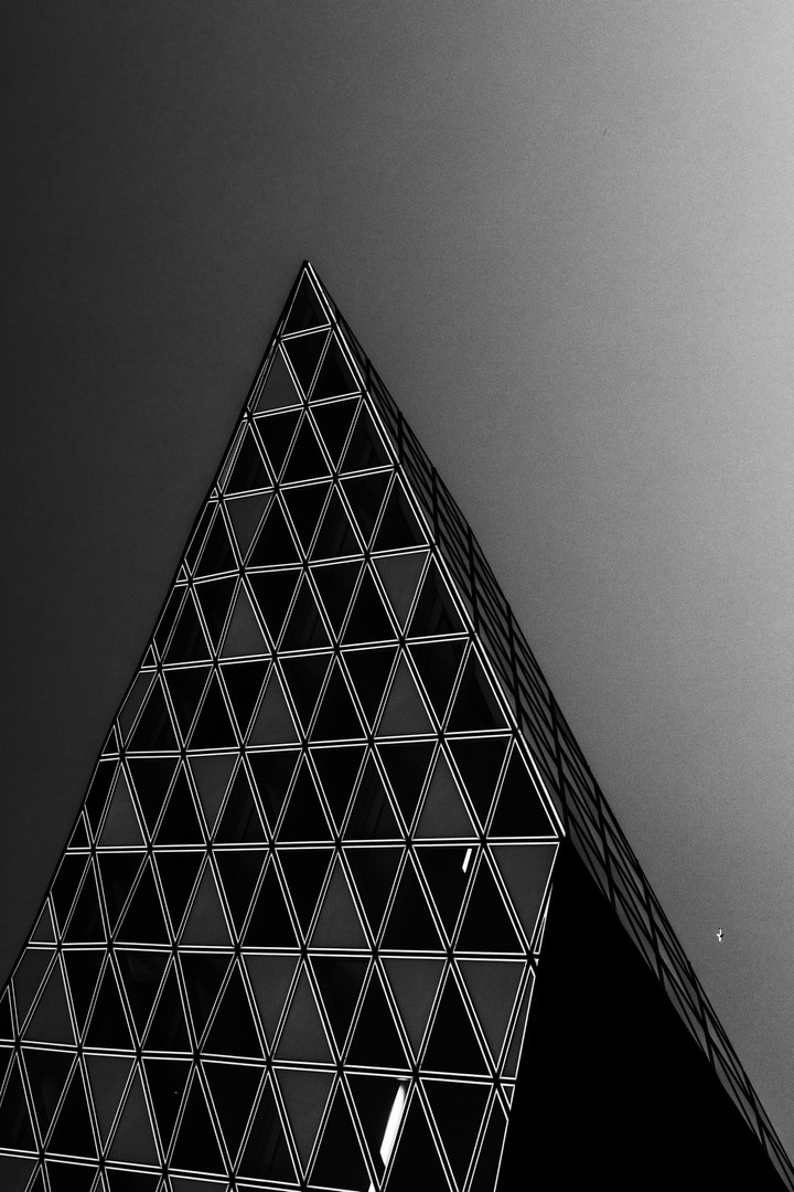 Dramatic high contrast close-up section from the glass extension of The Port House by Zaha Hadid, forming a sharp point and resembling a pyramid; to the right is a aeroplan mid-flight — Antwerp, Belgium