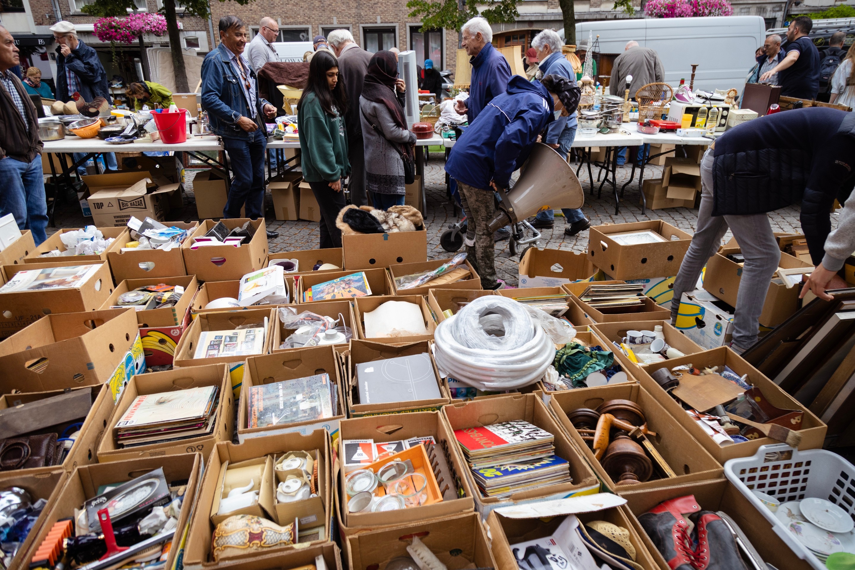 A street scene of a jumble sale and flower market, and in the foreground dozens of neatly lined-up boxes containing various knick-knack — Antwerp City, Belgium