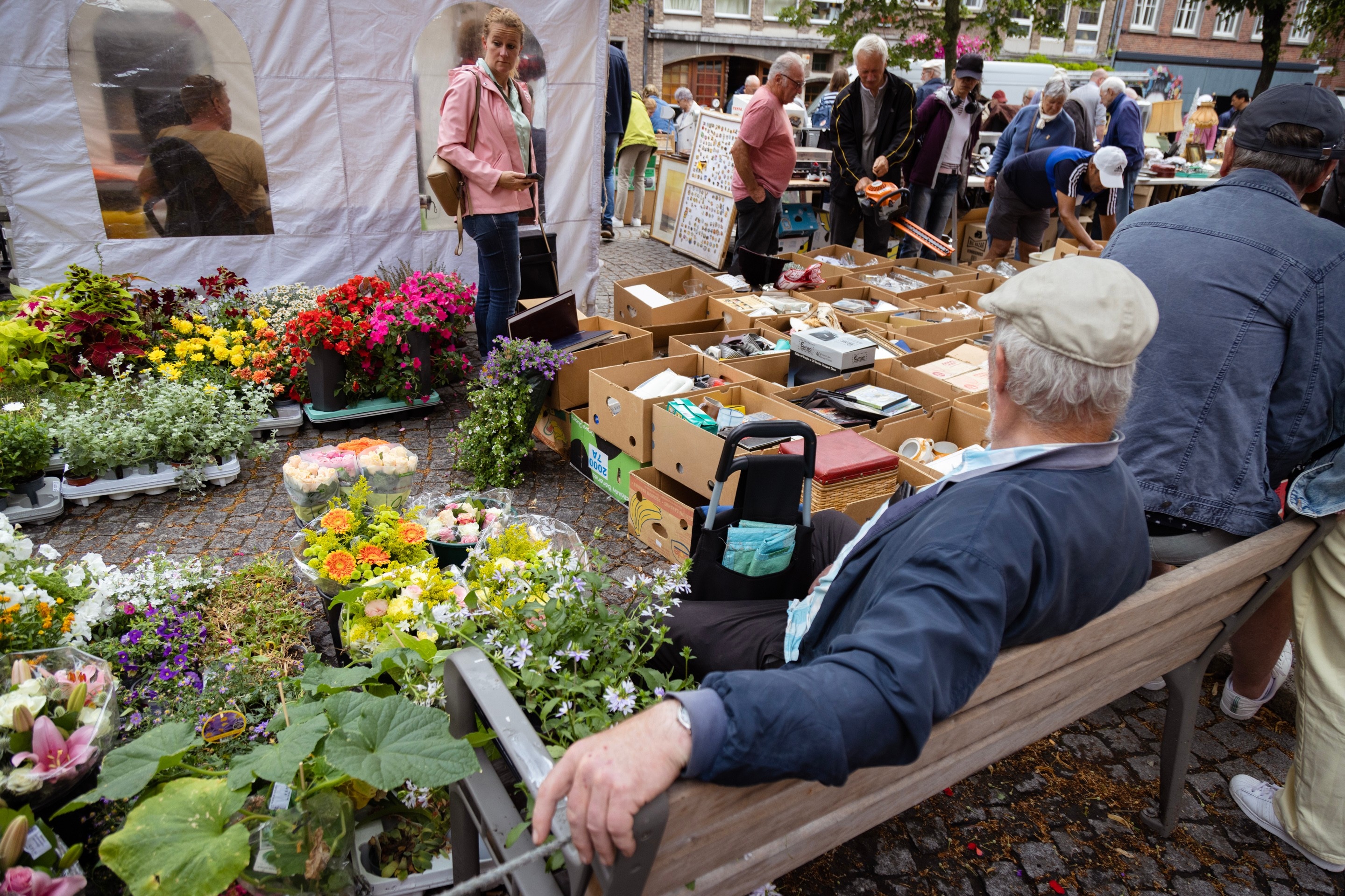 A man sits on a bench in the foreground and behind him a street scene of a jumble sale and flower market — Antwerp City, Belgium