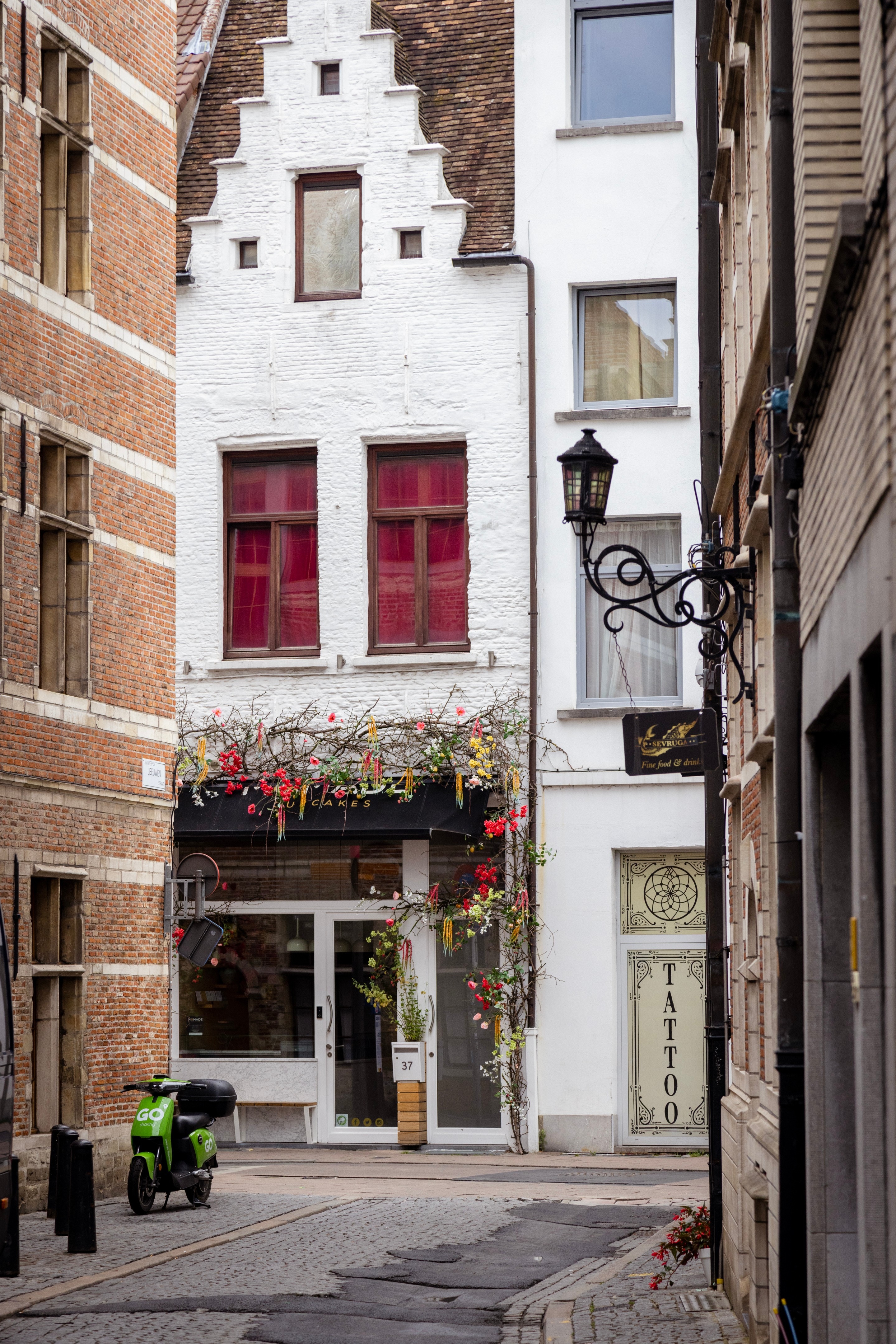 A cobbled street in Antwerp City, with one shop decorated with a lovely arrangement of drooping flowers. To the right there's an old iron street lamp with stained glass — Belgium