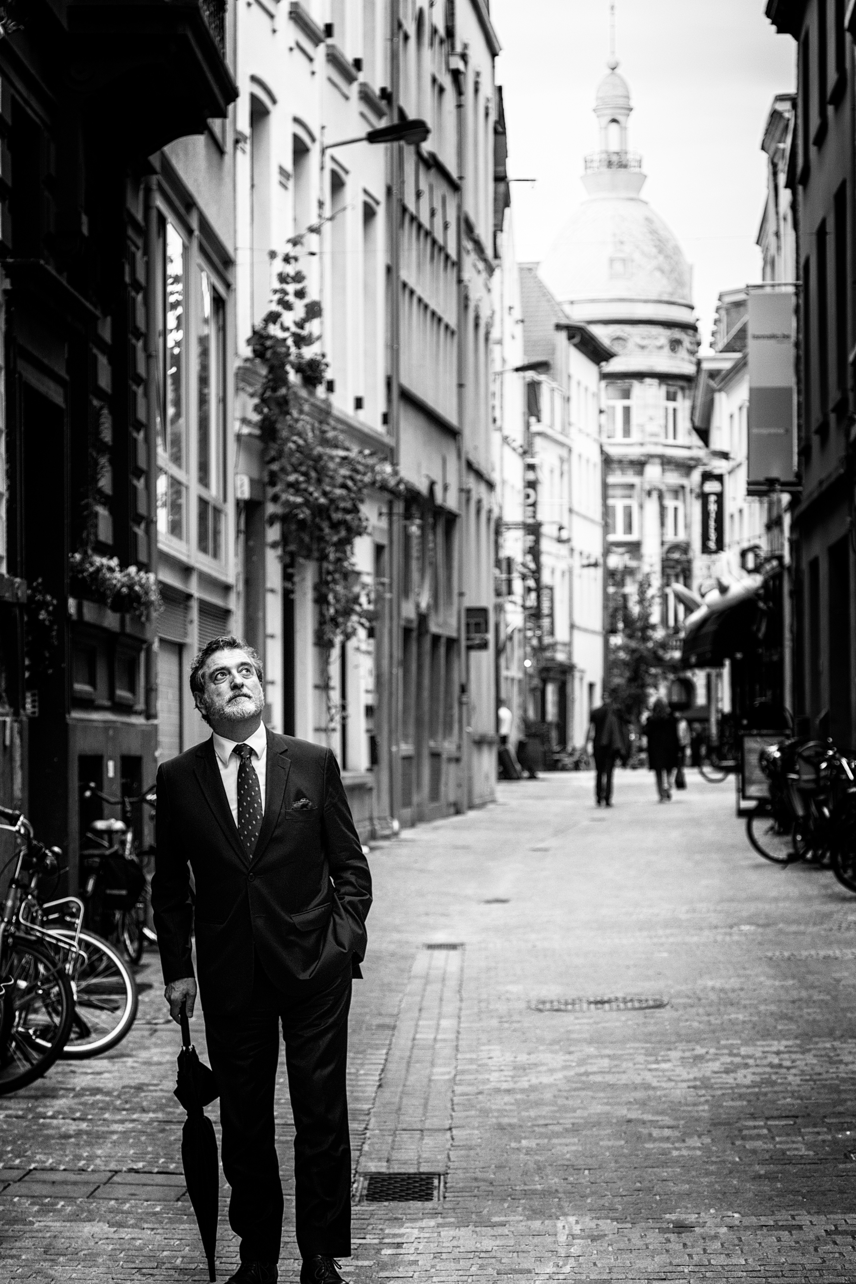 A street scene in black and white, with a prominant suited man, leaning on is folder umbrella, stands in the foreground looking up in the sky with a ponderous expression — Antwerp City, Belgium