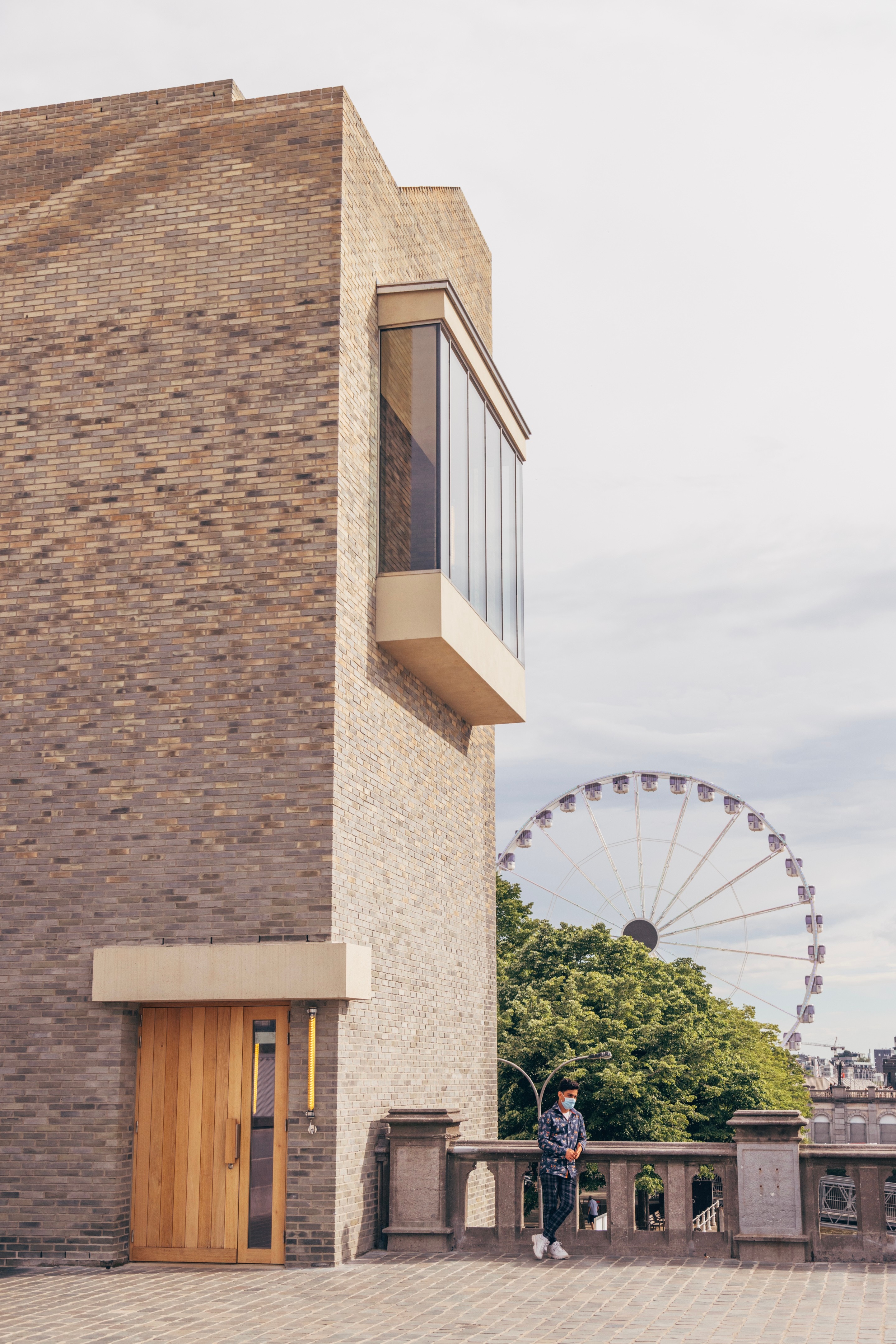 A man leans on a wall next to he new architectural tourist centre wing of the Het Steen fort, designed by Pieter Vermeersch. In the background, and behind the man is a large ferris wheel — Antwerp City, Belgium