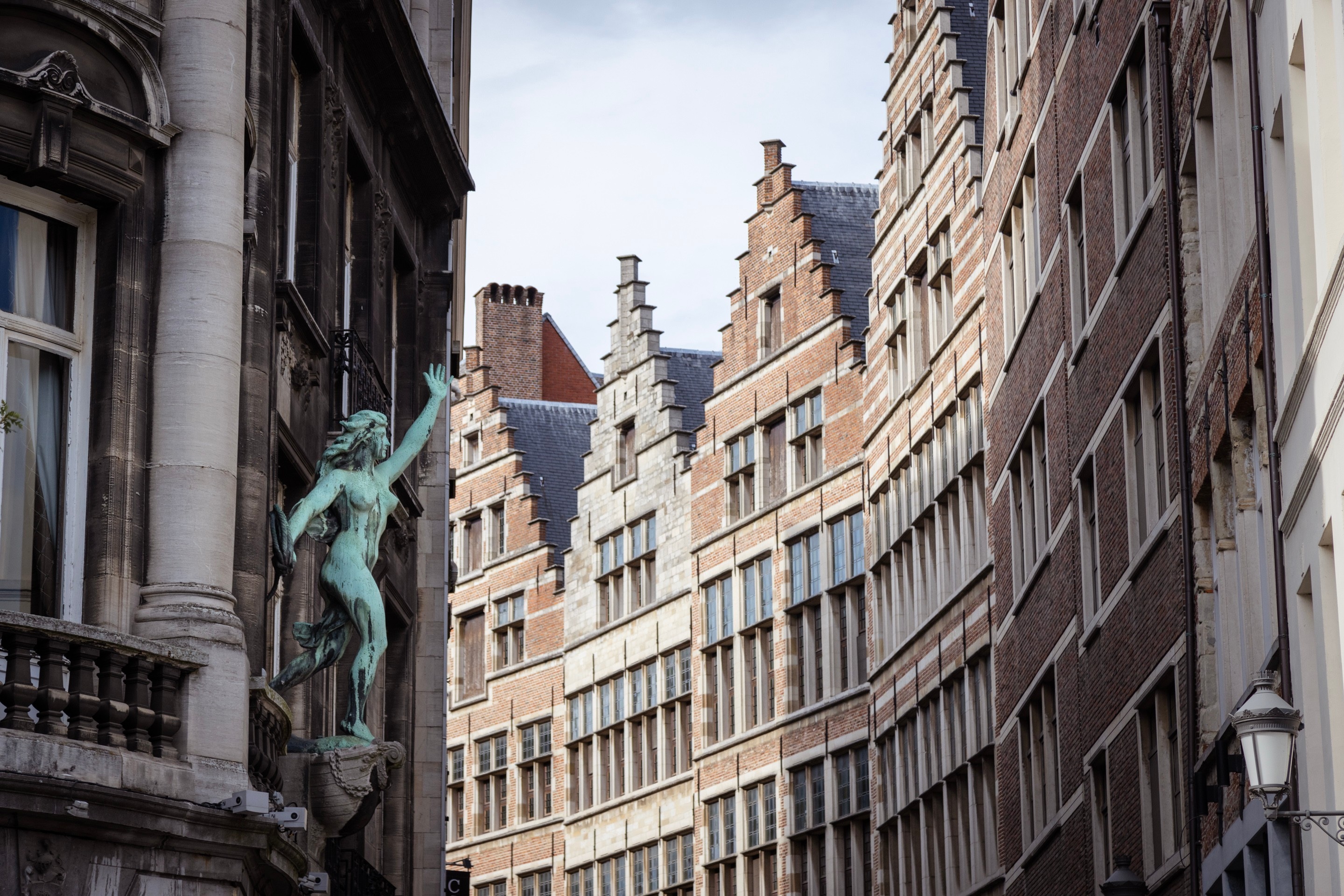 To the left is a bronze nude female statue throwing rope on a street off the, and to the right a line of mediaeval storefront buildings off the Main Square — Antwerp City, Belgium