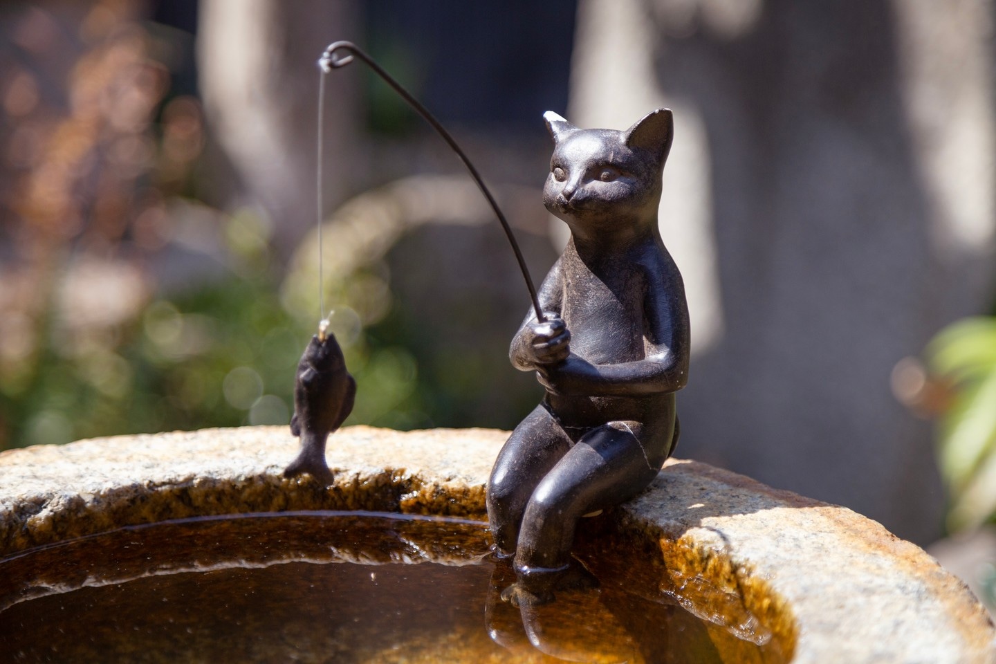 A bronze cat figure sits on a stone birdbath's rim holding a fishing rod, and a caught fish — Kurashiki Town, Okayama Prefecture, Japan
