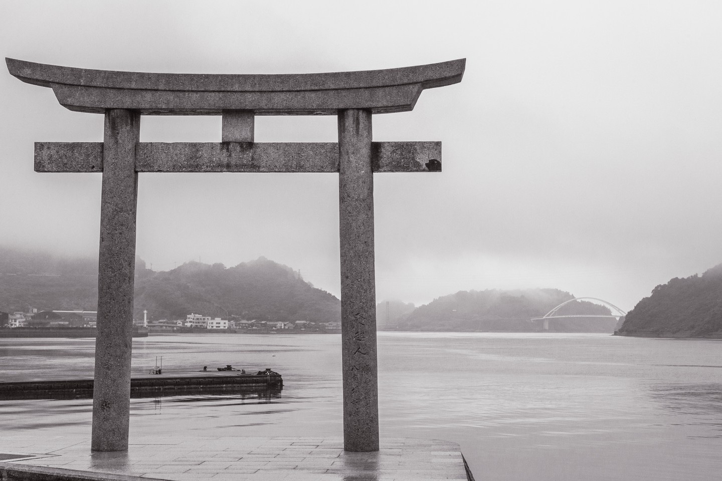 A stone Shinto Torii; Japanese spiritual gate with the ocean, islands and bridge spanning two islands seen the background in black and white — Mitarai village, Osaki Shimojima Island, Geiyo Region.