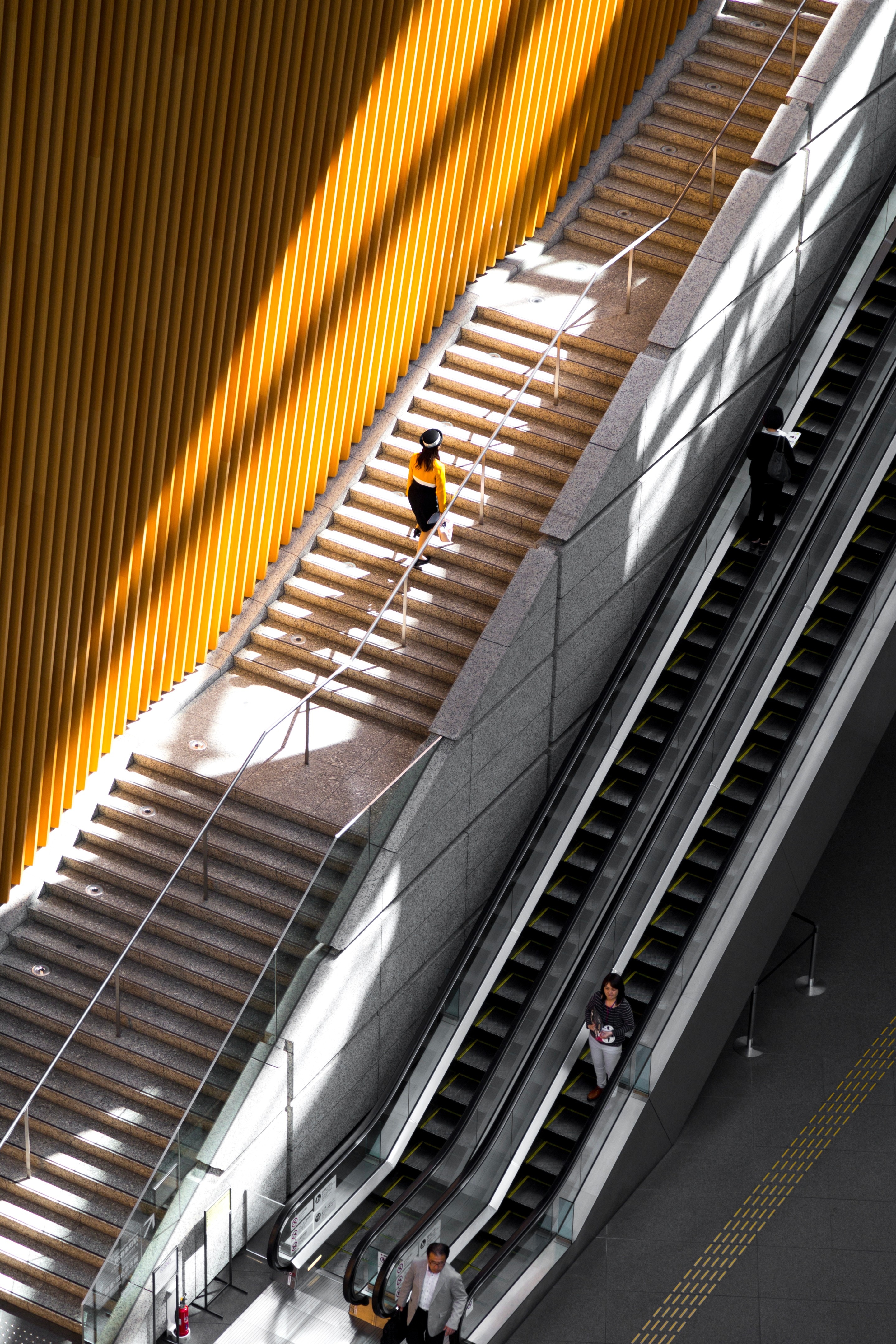 A woman in an orange top walks up a long concrete staircase, itself lined with matching orange walls. The sun both illuminates and casts long shadows creating striking contrast and interest. Taken at the Tokyo International Forum building — Marunouchi, Tokyo, Japan